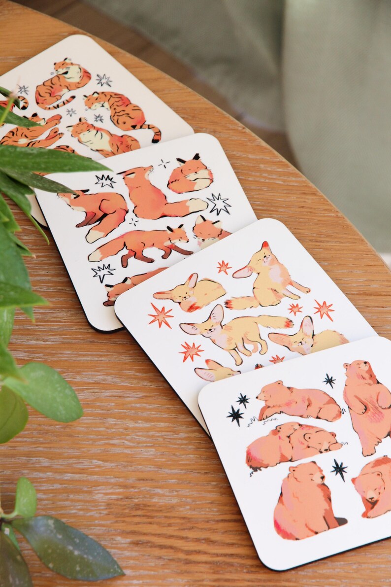 May include: Set of four square coasters featuring illustrations of various animals. The coasters showcase orange tigers, foxes, fennec foxes, and bears, with star and lightning bolt accents. The coasters are on a wooden surface.