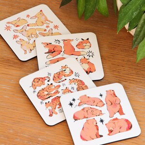 May include: Set of four square coasters with animal illustrations. Each coaster features a different animal: foxes, tigers, bears, and rabbits. The animals are in orange and pink, with star accents on a white background.
