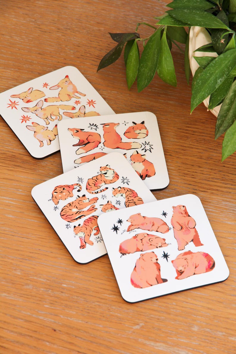 May include: Set of four square coasters with animal illustrations. Each coaster features a different animal: foxes, tigers, bears, and rabbits. The animals are in orange and pink, with star accents on a white background.