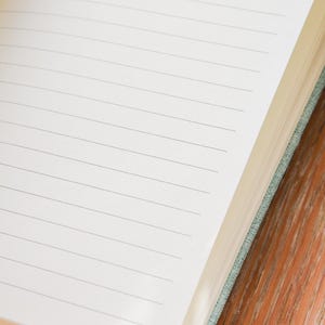 Puede incluir: Cuaderno abierto con p&aacute;ginas rayadas. Las p&aacute;ginas son blancas con finas l&iacute;neas grises. El cuaderno tiene una cubierta de tela verde azulado claro y un marcador de cinta verde azulado. El cuaderno est&aacute; sobre una superficie de madera.