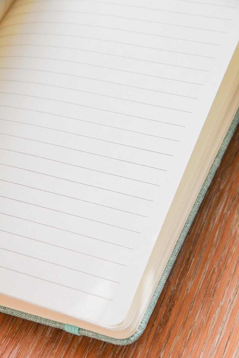 Puede incluir: Cuaderno abierto con p&aacute;ginas rayadas. Las p&aacute;ginas son blancas con finas l&iacute;neas grises. El cuaderno tiene una cubierta de tela verde azulado claro y un marcador de cinta verde azulado. El cuaderno est&aacute; sobre una superficie de madera.