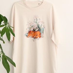 May include: Cream-colored t-shirt featuring a watercolor illustration of a sleeping orange tiger amidst white flowers, green leaves, and stars. The design is centered on the front of the shirt, creating a whimsical and artistic aesthetic.