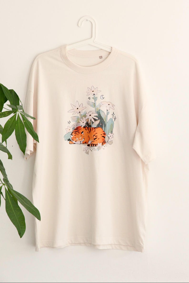 May include: Cream-colored t-shirt featuring a watercolor illustration of a sleeping orange tiger amidst white flowers, green leaves, and stars. The design is centered on the front of the shirt, creating a whimsical and artistic aesthetic.