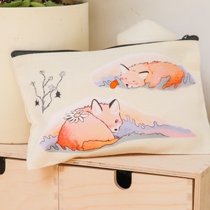 Może przedstawiać: Biała płócienna kosmetyczka z czarnym zamkiem błyskawicznym. Kosmetyczka przedstawia ręcznie rysowaną ilustrację dwóch śpiących lisów na pastelowym tle. Jeden lis ma stokrotkę na grzbiecie.