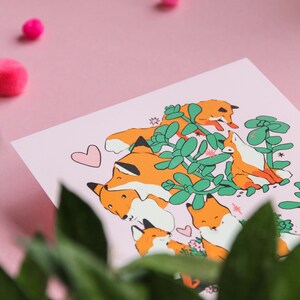May include: A pink background with a white card featuring a group of orange foxes with black eyes and noses. The foxes are surrounded by green leaves and flowers. The card has a pink heart in the top left corner.