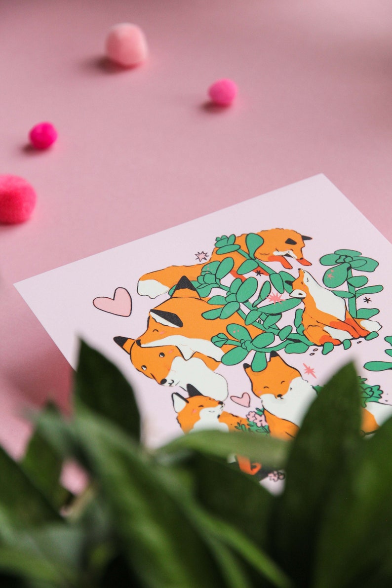 May include: A pink background with a white card featuring a group of orange foxes with black eyes and noses. The foxes are surrounded by green leaves and flowers. The card has a pink heart in the top left corner.