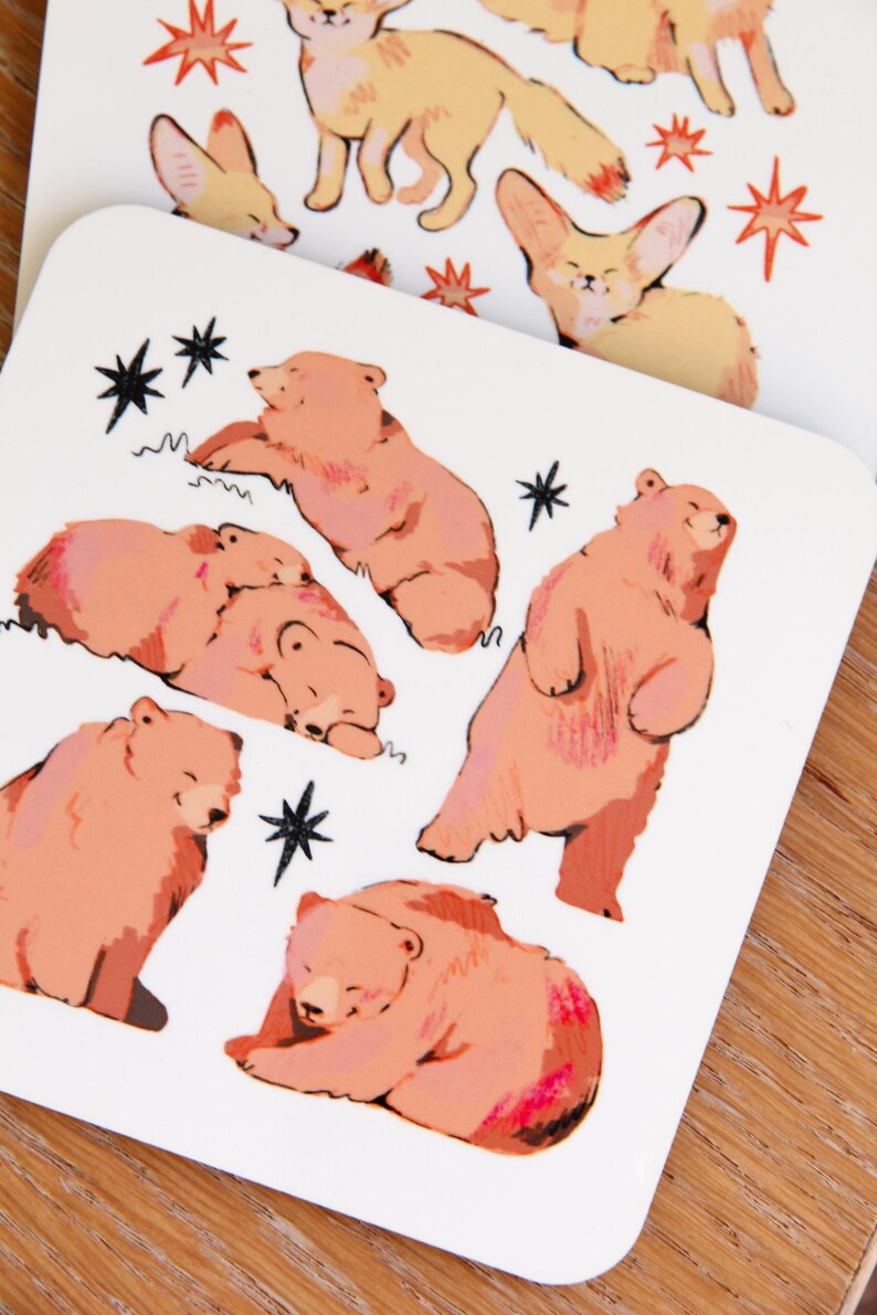 May include: Two white square cards with illustrations of animals. One card features foxes and stars in orange and yellow. The other card has pink bears and black stars. The cards are on a wooden surface.