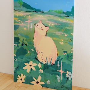 May include: Art print featuring a light-colored cat basking in a field of flowers under a blue sky. The illustration has a whimsical style with soft colors and highlights. The artist's signature "MADI" is visible in the lower left corner.