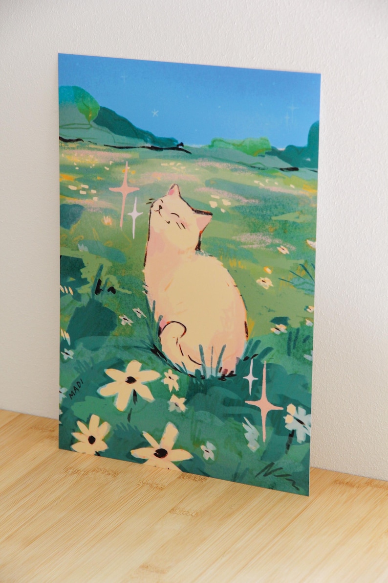 May include: Art print featuring a light-colored cat basking in a field of flowers under a blue sky. The illustration has a whimsical style with soft colors and highlights. The artist's signature "MADI" is visible in the lower left corner.