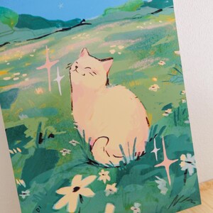 May include: A whimsical illustration of a cream-colored cat sitting in a field of green grass and white flowers. The cat is smiling with its eyes closed. The background features a blue sky and rolling hills. The artist's name, MADI, is in the lower left corner.