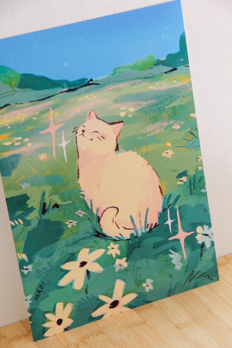 May include: A whimsical illustration of a cream-colored cat sitting in a field of green grass and white flowers. The cat is smiling with its eyes closed. The background features a blue sky and rolling hills. The artist's name, MADI, is in the lower left corner.