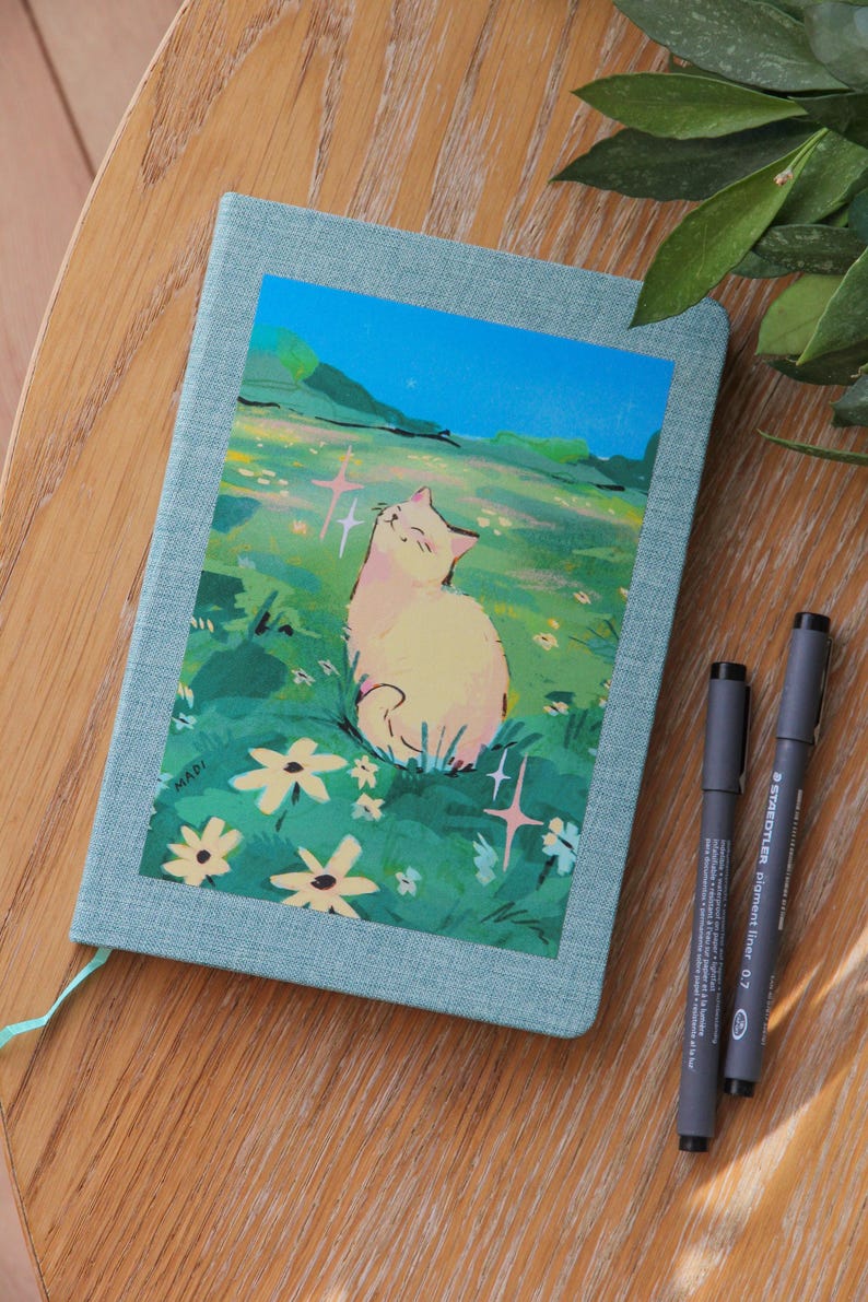 Puede incluir: Un diario azul claro con una ilustraci&oacute;n de un gato en la portada. La ilustraci&oacute;n representa un gato en un campo de flores. Dos bol&iacute;grafos negros est&aacute;n colocados sobre la superficie de madera junto al diario.