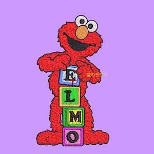 Op de afbeelding: Geborduurde Elmo-figuur met blokken met de tekst "ELMO" op een paarse achtergrond. De rode Elmo heeft een zwarte glimlach, witte ogen en een oranje neus. De blokken zijn blauw, groen, geel en paars.