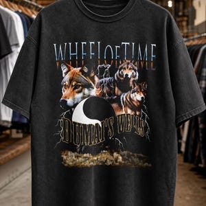 May include: A black short-sleeve t-shirt featuring a graphic print with the text WHEEL OF TIME and DUMAI'S WELLS. The design includes three wolves, a yin-yang symbol, and lightning bolts against a dark background, printed on the front of the shirt.