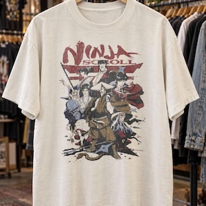 May include: A cream-colored short-sleeve t-shirt featuring a graphic print of the anime Ninja Scroll. The design displays multiple characters in a battle scene with the red text Ninja Scroll at the top, set against a dark red torii gate background.