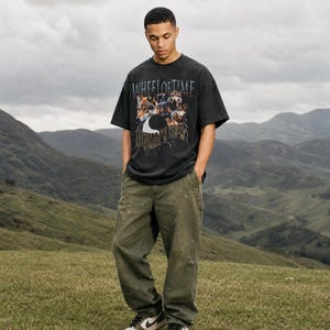 May include: A black short-sleeve t-shirt featuring a graphic print of wolves and the text "WHEEL OF TIME" and "HUMAN'S WILL" paired with olive green loose-fit pants and black and white sneakers. The shirt has a crew neckline and a relaxed fit.
