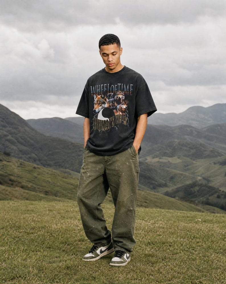 May include: A black short-sleeve t-shirt featuring a graphic print of wolves and the text "WHEEL OF TIME" and "HUMAN'S WILL" paired with olive green loose-fit pants and black and white sneakers. The shirt has a crew neckline and a relaxed fit.