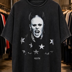 May include: A charcoal gray short-sleeve t-shirt featuring a black and white graphic print of a person with spiky hair and an open mouth expression. The design includes several white stars surrounding the portrait and the name KEITH printed below.