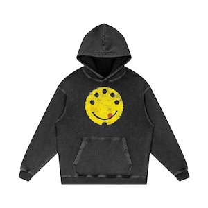 Puede incluir: Una sudadera con capucha negra con un gran smiley amarillo en la parte delantera. El smiley tiene seis puntos negros y una lengua roja. La sudadera tiene un bolsillo delantero y una capucha con cordón.