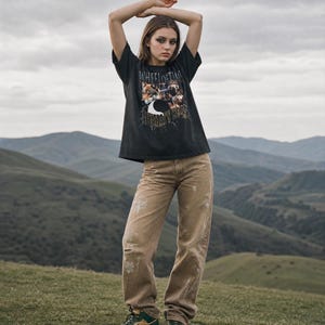 May include: A black short-sleeve t-shirt featuring a graphic print of wolves and the text "WHEEL OF TIME" and "THE DRAGON REBORN". The shirt is paired with tan distressed denim jeans that have light-colored paint splatters and a single knee rip.