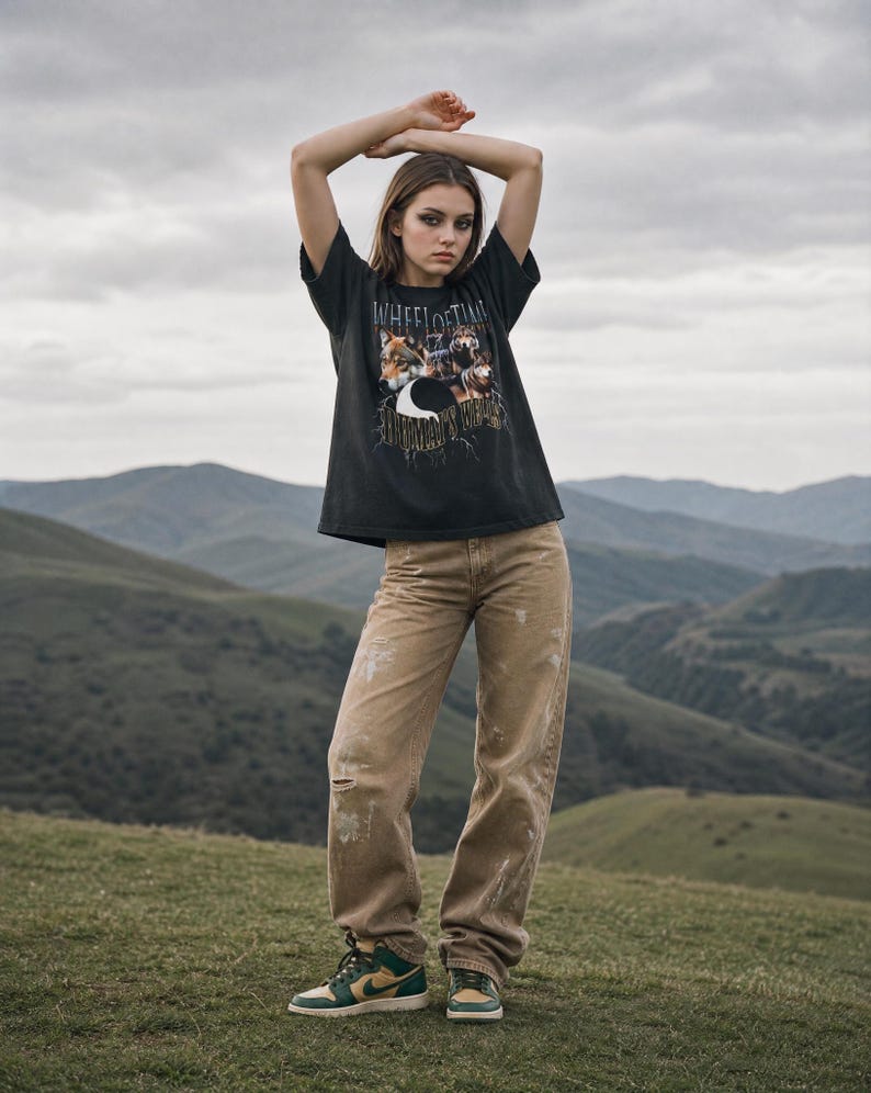 May include: A black short-sleeve t-shirt featuring a graphic print of wolves and the text "WHEEL OF TIME" and "THE DRAGON REBORN". The shirt is paired with tan distressed denim jeans that have light-colored paint splatters and a single knee rip.