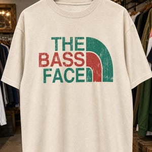 Peut inclure: Un t-shirt beige à manches courtes avec une impression graphique vieillie sur la poitrine. Le texte THE BASS FACE est écrit en lettres vertes et rouges, accompagné d'un motif graphique en quart de cercle vert et rouge sur la droite.
