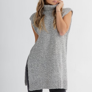 May include: A gray and white knit turtleneck sweater with a high neck and sleeveless design. The sweater has side slits and a long, loose fit.