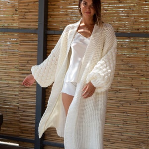 May include: A white knitted cardigan with a long, open front and large, puffy sleeves. The cardigan is made of a soft, textured yarn and has a relaxed fit.
