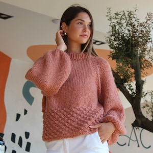 May include: A soft, pink knitted sweater with a slightly oversized fit and a textured design on the bottom. The sweater has long sleeves with a balloon-like shape.