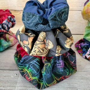 May include: A stack of three fabric drawstring bags. The top bag is navy blue with a white pattern. The middle bag is black with a white and brown mushroom pattern. The bottom bag is black with a rainbow and black insect pattern.
