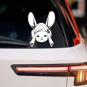 Può includere: Adesivo in vinile bianco di un coniglio dei cartoni animati con un cappello, orecchie grandi e un'espressione carina. L'adesivo è applicato sul lunotto posteriore di un'auto bianca.
