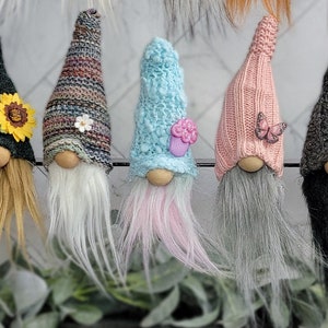 May include: Five gnomes with different colored hats and beards. The gnomes are wearing hats with various embellishments, including a sunflower, a butterfly, a flower, and a cupcake.