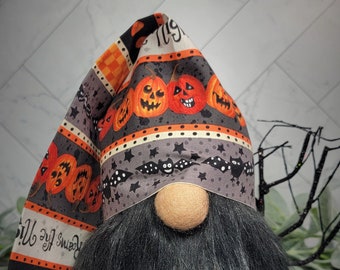 May include: A Halloween gnome with a black beard and a hat with a pattern of pumpkins, bats, and stars. The hat has the text "Boo!" on it. The gnome is holding a black wooden bat.