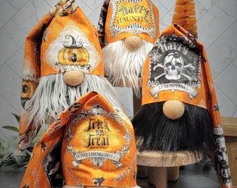 May include: Four Halloween gnomes with orange and white patterned hats. The hats feature text that says "Happy Haunting" and "Trick or Treat". The gnomes have white, gray, and black beards and tan noses. One gnome has a small orange tree.