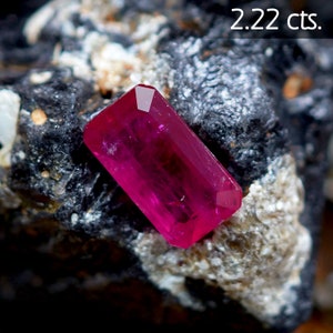 May include: A vibrant pink ruby gemstone, weighing 2.22 carats, is displayed on a dark grey rock. The ruby has a rectangular shape with facets that catch the light.