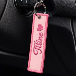 Peut inclure: Porte-clés brodé rose avec le nom "Titine" et un motif cœur. Le porte-clés rectangulaire a une bordure rose et est attaché à un porte-clés argenté. Le porte-clés est près du contact d'une voiture.