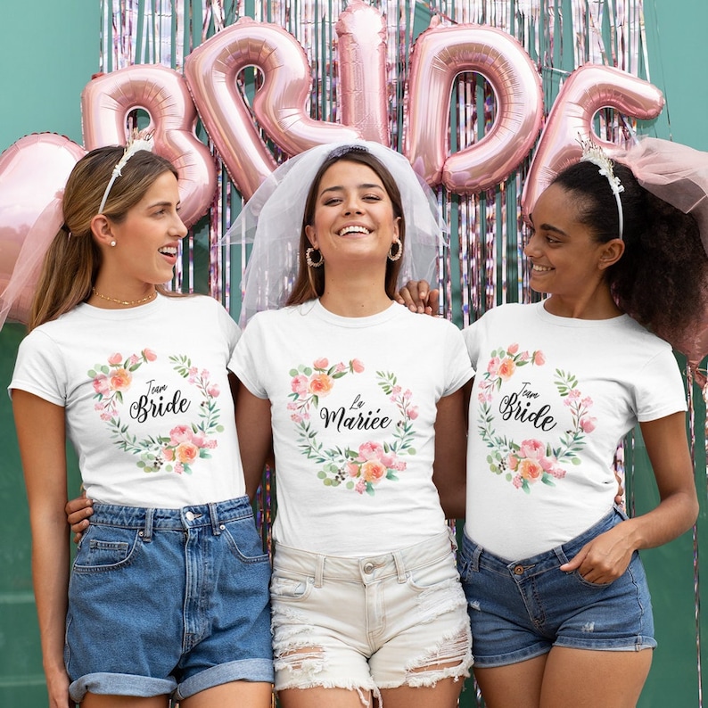 May include: Three women wearing white t-shirts with floral wreaths and the text "Team Bride" and "La Mari&eacute;e". The women are smiling and standing in front of a teal wall with pink balloons that spell out "Bride".