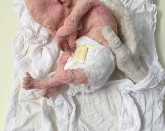 May include: A small, pink, felt baby doll wearing a white diaper with a yellow pad. The doll is lying on a white cloth with a hand made of felt holding it.