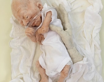 May include: A small, pale, bald baby doll wearing a white onesie and a clear breathing tube lies on a white cloth. The doll's eyes are closed and its mouth is slightly open. The doll's limbs are thin and its fingers are curled.