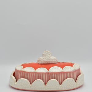 May include: A ceramic cake-shaped container with a coral-colored top and a white cloud-shaped handle. The sides are pink with vertical white stripes. The base and top edges have a scalloped design with dotted texture.