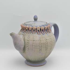 May include: A handcrafted ceramic teapot with a unique design. The teapot features a light purple handle, spout, and base, with a textured body in cream and light blue. The lid is topped with a small knob. The rim is decorated with a series of tooth-like shapes.