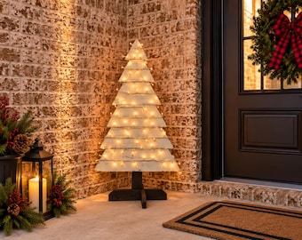 Puede incluir: Un árbol de Navidad decorativo de madera con tablones horizontales de color blanco desgastado apilados en forma triangular. El árbol está iluminado por una cadena de luces blancas cálidas y se sostiene sobre una base de madera en cruz.
