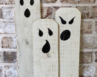 Puede incluir: Tres decoraciones de fantasmas de madera blanca con caras pintadas de negro. Los fantasmas son de diferentes tamaños y tienen diferentes expresiones faciales.