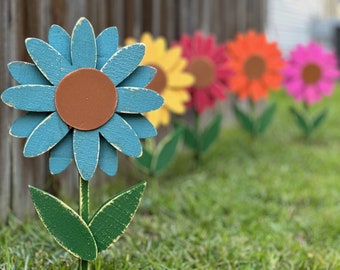 May include: A row of wooden flower decorations in a garden setting. The flowers are painted in various colours, including blue, yellow, red, and pink. The flowers are arranged in a line, with the blue flower in the foreground.