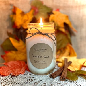 Peut inclure: Une bougie allumée en cire de soja dans un bocal Mason avec une étiquette marron indiquant "Pumpkin Spice". La bougie est attachée avec de la ficelle et entourée de feuilles d'automne et de bâtons de cannelle. Le texte "THE LITTLE ARTISAN" et "Handmade in Canada" sont également visibles.