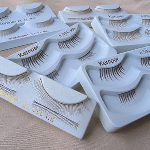 May include: Several sets of false eyelashes are displayed on a light beige surface. Each set is packaged in a white plastic tray with the brand name "Kemper" printed in gold. The eyelashes are dark brown and appear to be new.