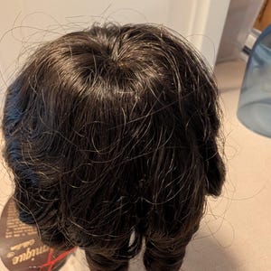 May include: A dark brown wig with a layered, wavy texture and a visible crown area. The hair is styled in loose, voluminous curls that fall around the head. A small, circular product tag with the text "unique collection" is attached to the side.