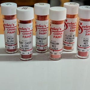 May include: A collection of six small glass bottles of Seeley's China Paint. Each bottle has a white cap and a label with the brand name and paint colour, including Cheek Blush, Ruby Cheek Blush, and Persian Red. The bottles contain various shades of red paint.