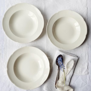 May include: Three off-white soup bowls with scalloped edges are arranged on a white tablecloth. A silver spoon with blue flowers and a white lace ribbon rests on a white linen napkin. The bowls are made of ceramic.