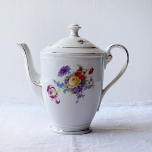 以下が含まれることがあります： 金色の縁取りと花柄の白い磁器製のコーヒーポット。花は黄色、赤、紫、青です。